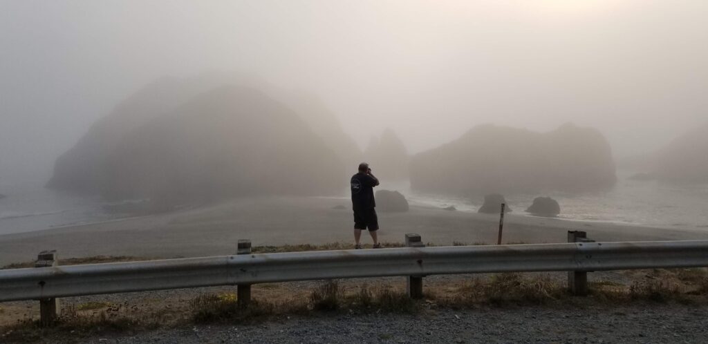 photo of a man taking a picture with a digital camera. he is standing on a beach in the northwest where it is cold and foggy. there are large rocks just off shore. 